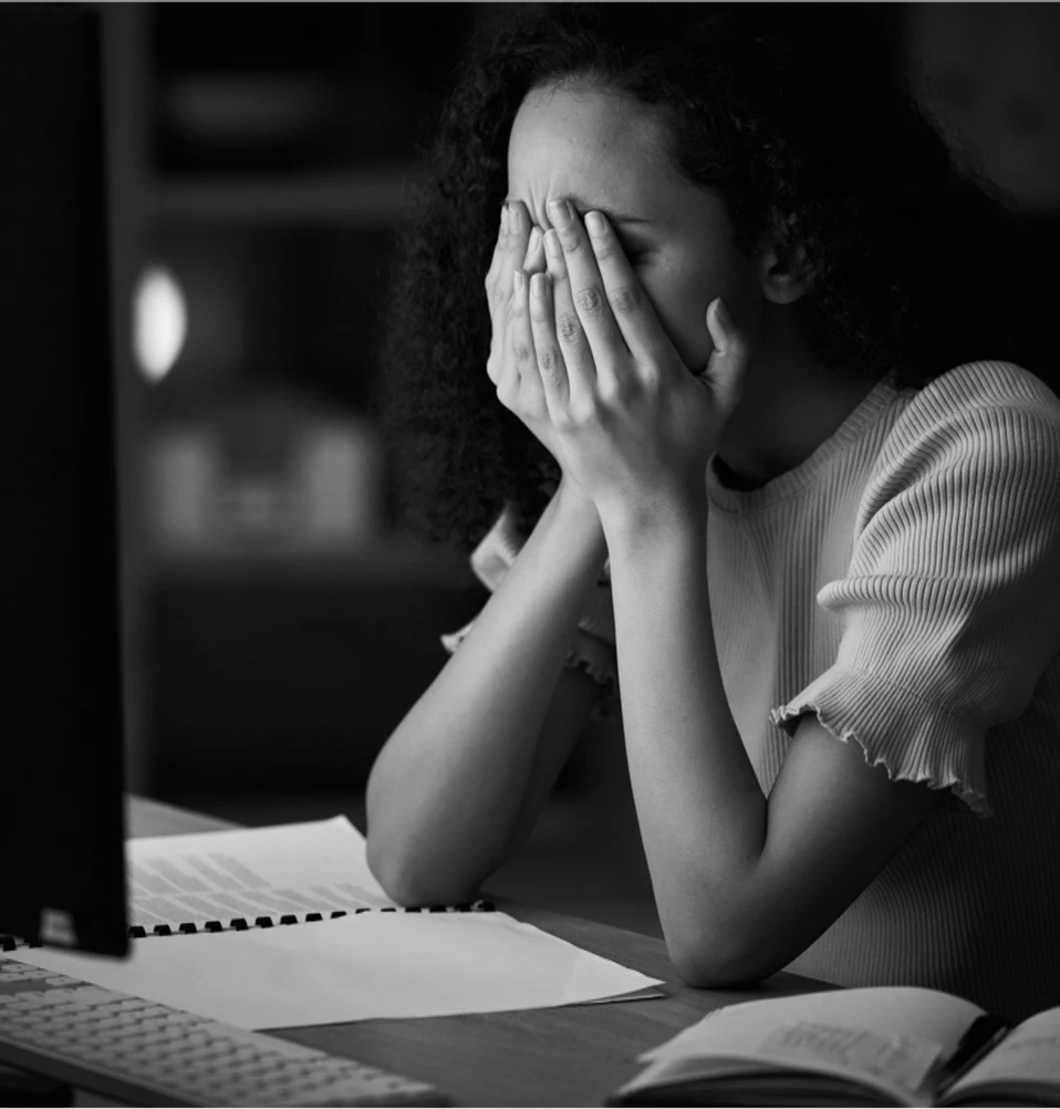Concurseira exausta com as mãos no rosto diante do computador e dos livros de estudo